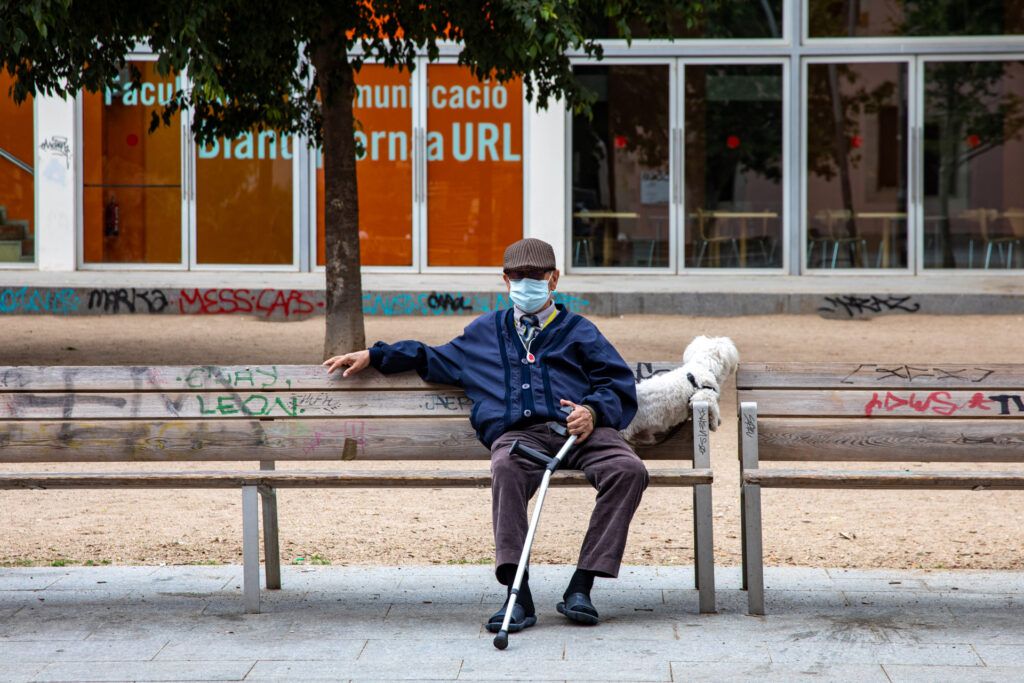 Hombre mayor con mascarilla y bastón sentado en un banco de la calle