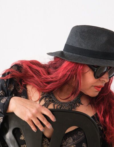 Mujer de cabello rojo con sombrero negro y gafas de sol inclinada en un retrato de estudio