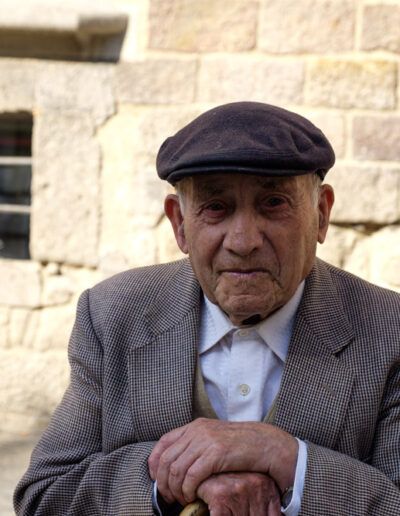 Retrato de un hombre mayor con gorra y traje junto a un muro de piedra en Barcelona