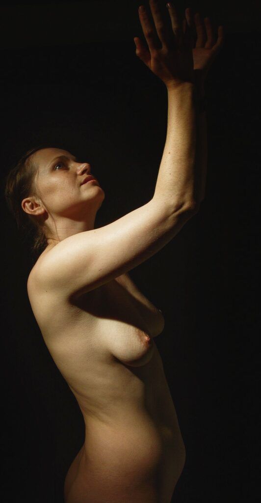 Desnudo artístico de una mujer de perfil con los brazos alzados y la mirada hacia arriba