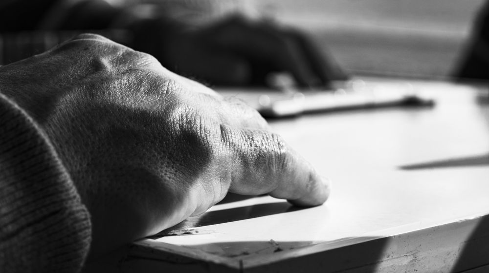 Mano de un jugador mayor apoyada sobre la mesa de dominó, en blanco y negro
