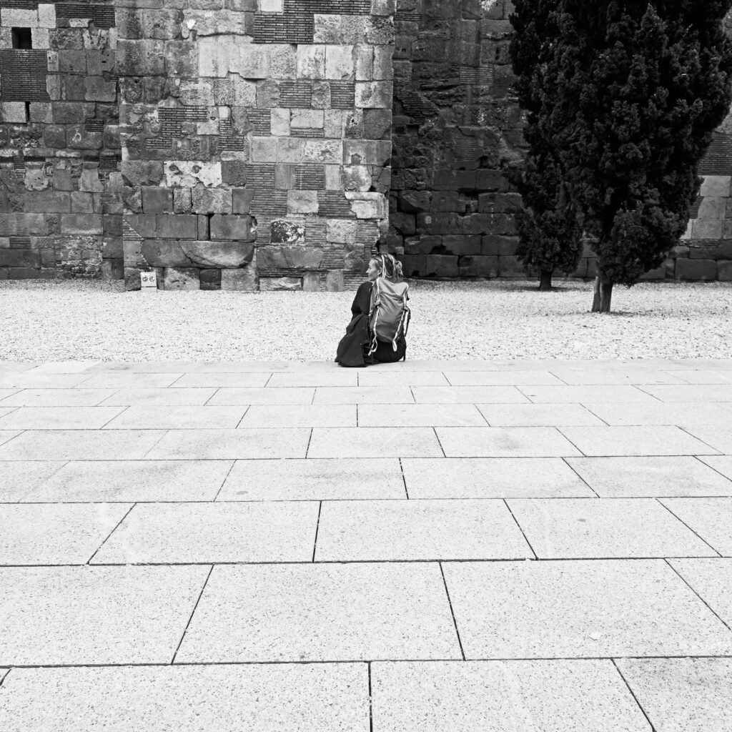 Persona sentada sola en una plaza de piedra junto a una antigua muralla, fotografía callejera en blanco y negro