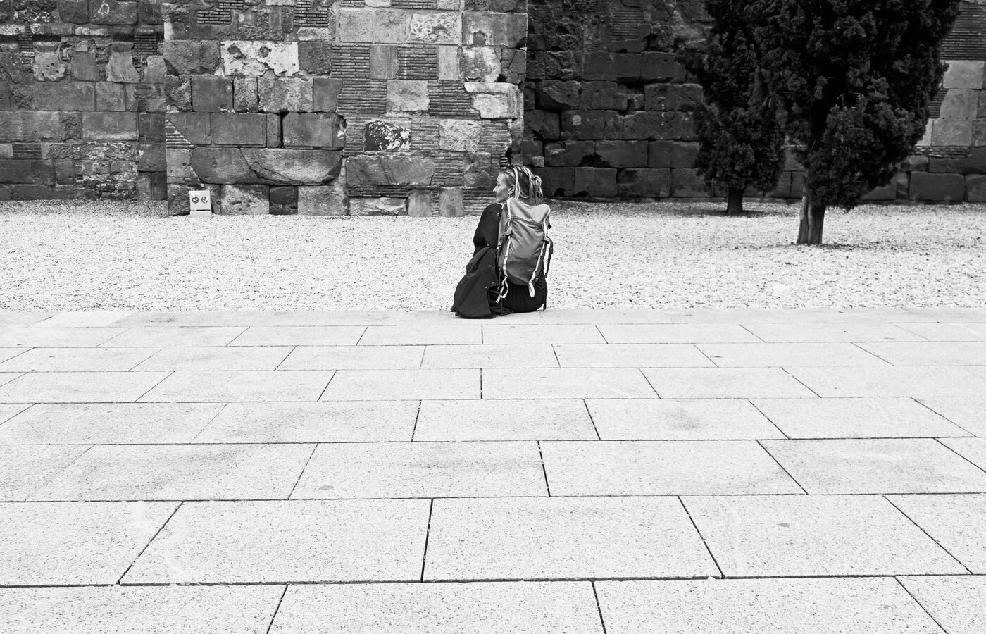 Persona sentada sola en una plaza de piedra junto a una antigua muralla, fotografía callejera en blanco y negro