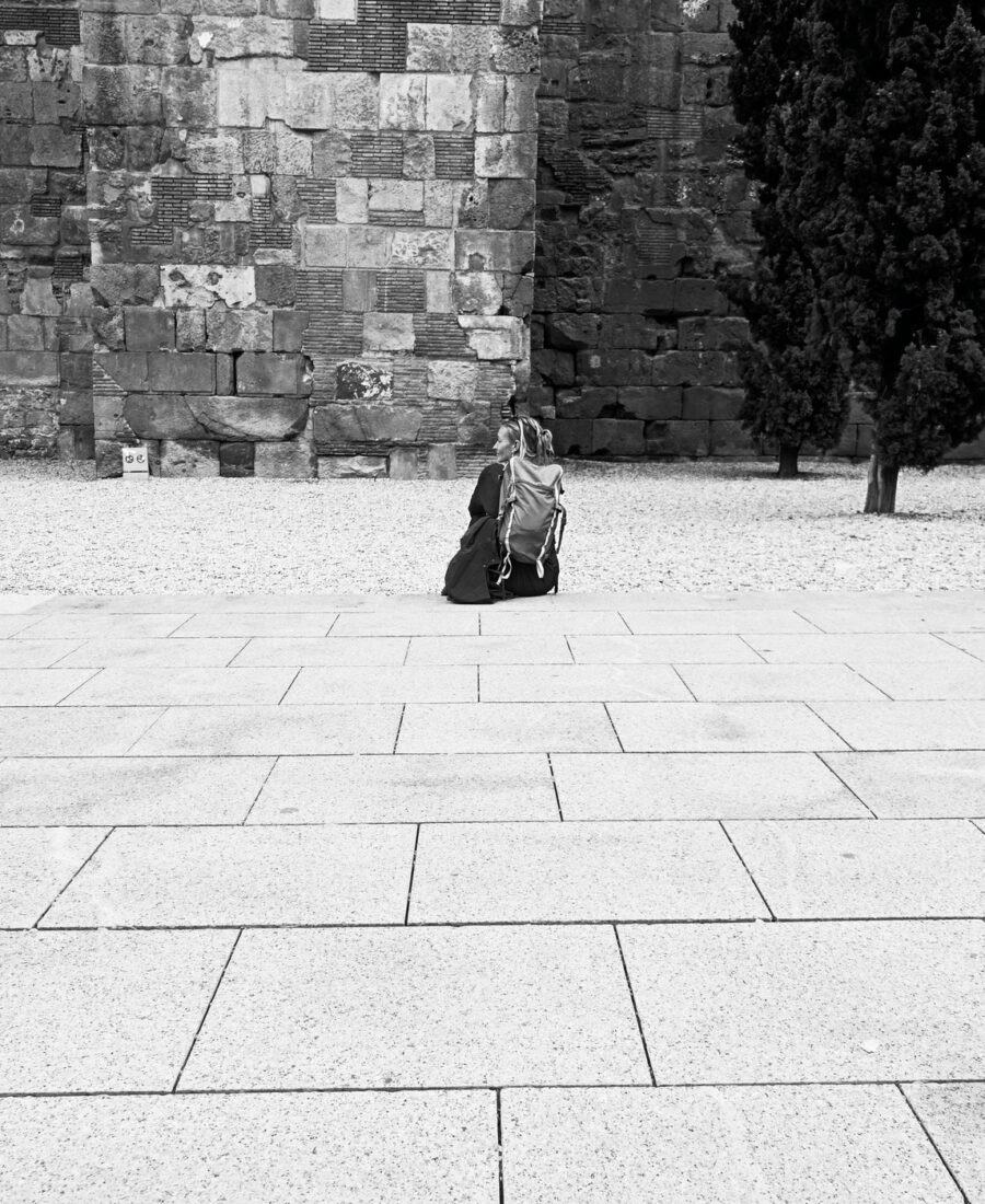 Persona sentada sola en una plaza de piedra junto a una antigua muralla, fotografía callejera en blanco y negro