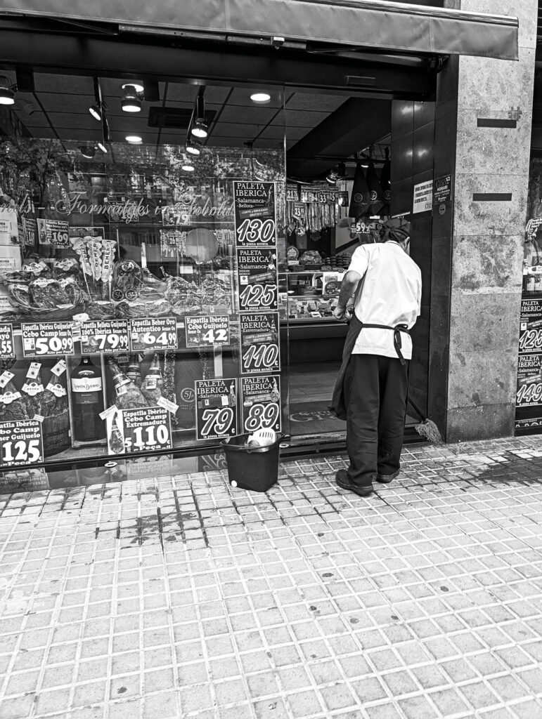 Dependiente limpiando la entrada de una tienda llena de carteles de precios, fotografía callejera en blanco y negro