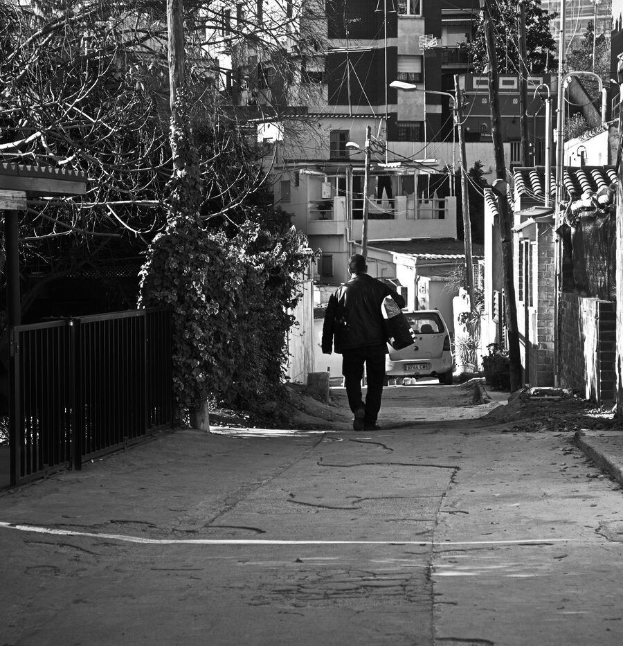 Hombre caminando de espaldas por una calle estrecha con árboles y luz dura, en blanco y negro