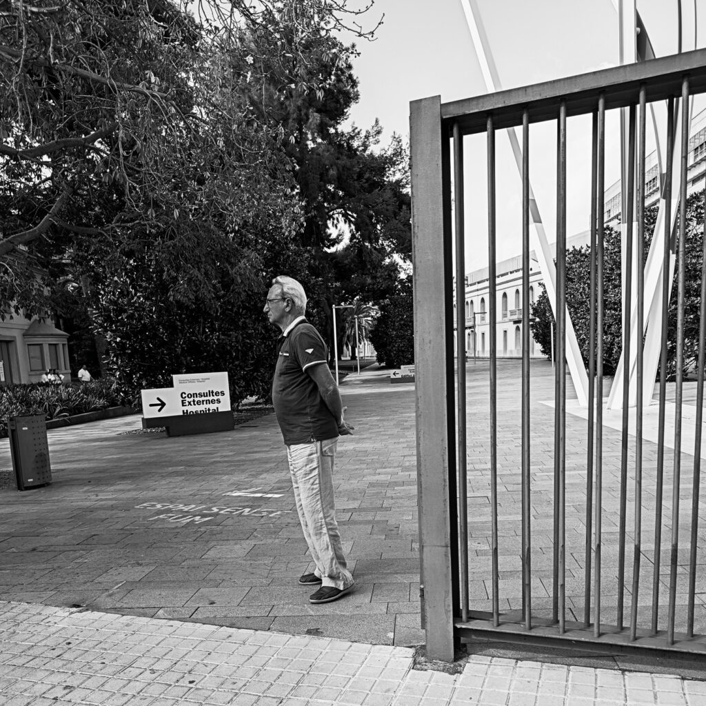 Hombre mayor de pie solo junto a una verja abierta en una plaza, fotografía en blanco y negro