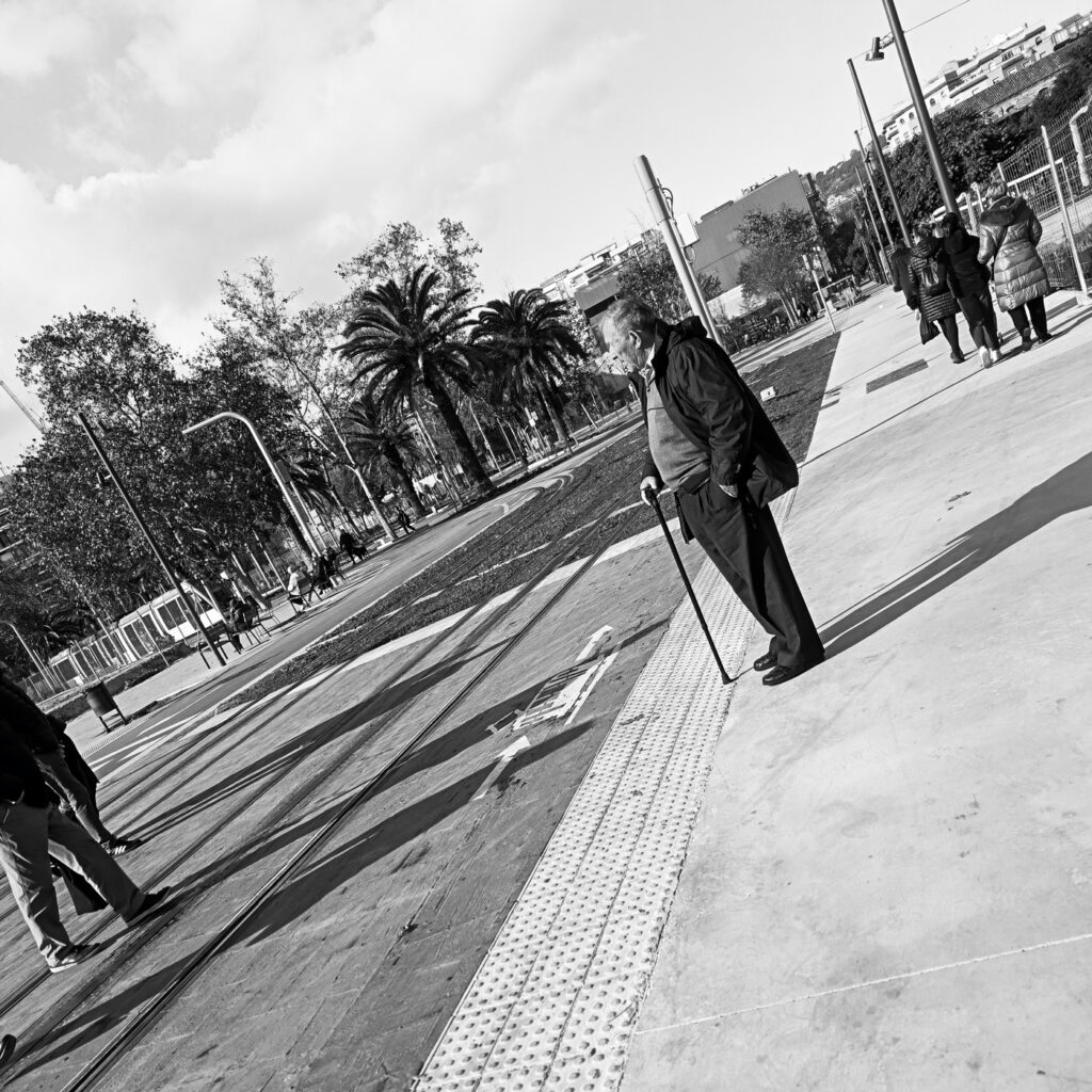 Hombre mayor con bastón caminando solo por una calle con palmeras, fotografía callejera en blanco y negro