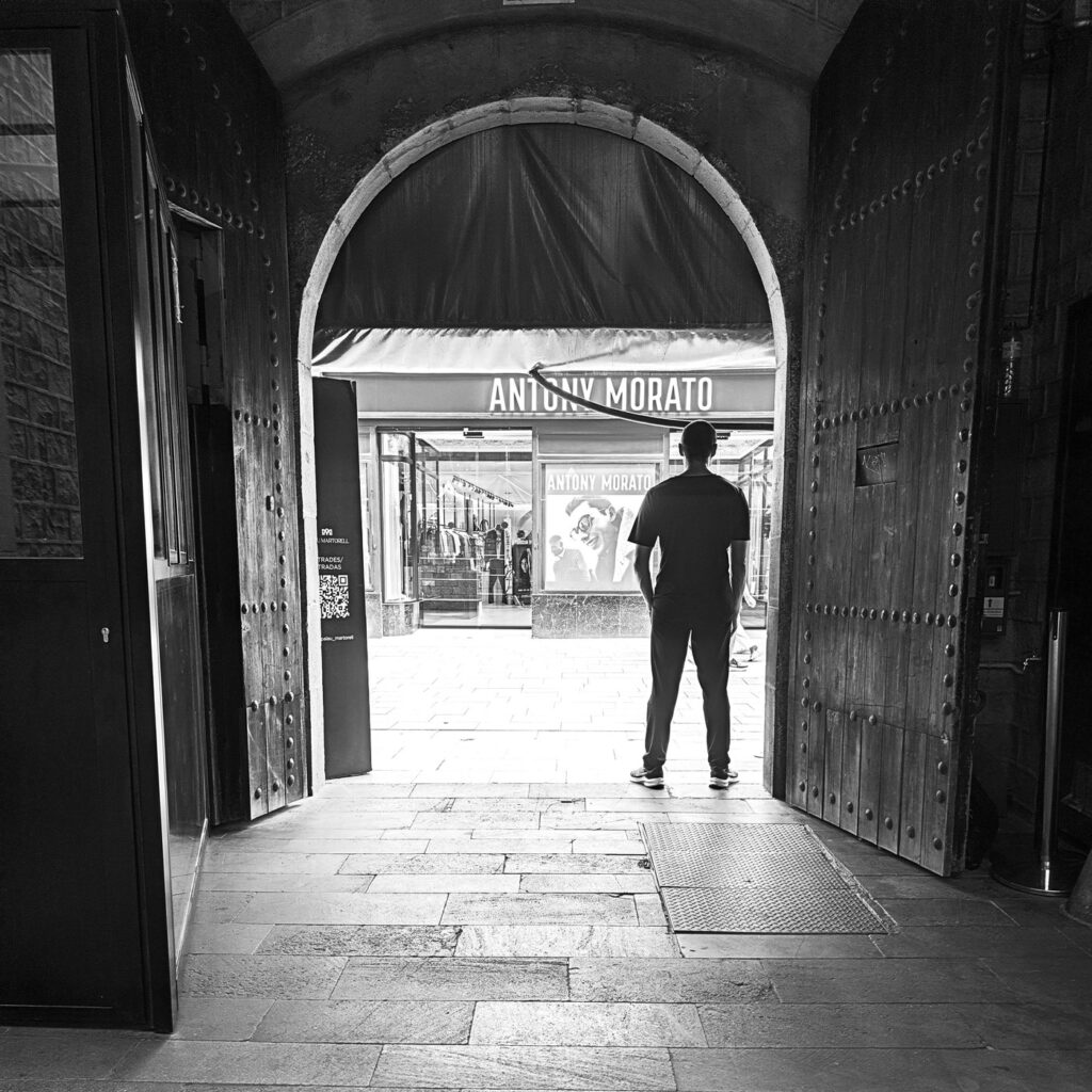Hombre de espaldas en la penumbra de un portal, mirando hacia la calle iluminada. Fotografía callejera en blanco y negro sobre la soledad